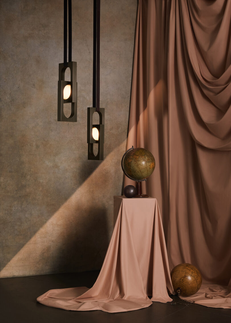 Interior featuring draped beige curtains, a table with matching cloth, two vintage globes, and modern hanging lamps against a textured wall.