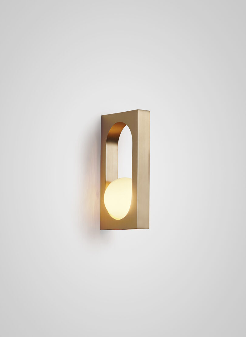A modern wall-mounted light fixture with an gold finish and a unique geometric design, casting a soft glow on a light grey wall.