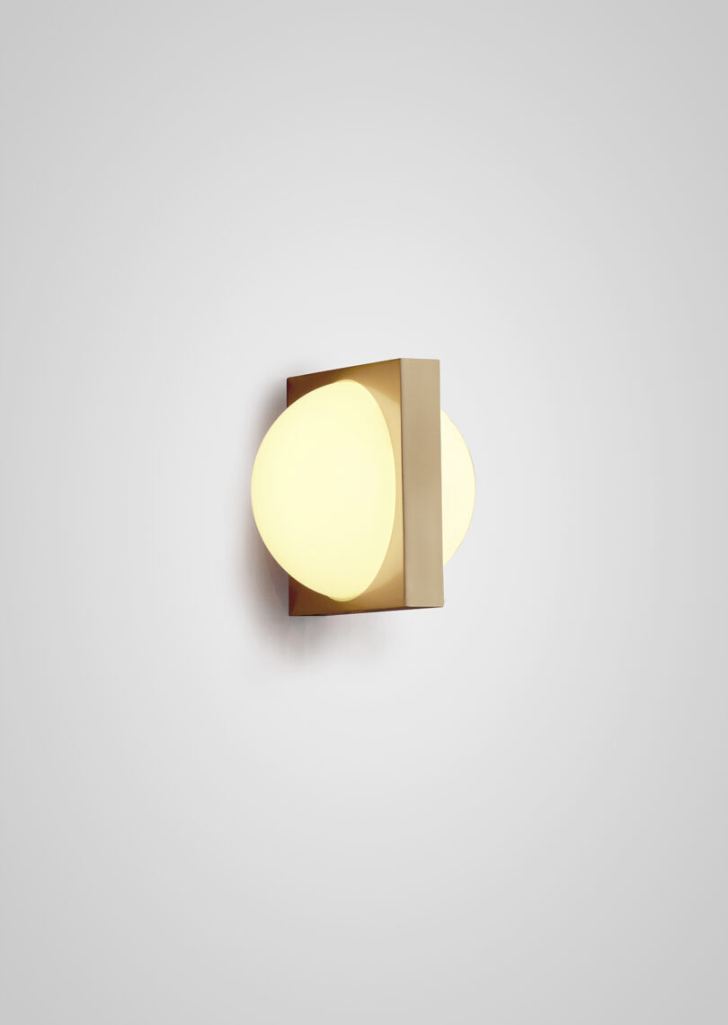 A modern wall-mounted light fixture with an gold finish and a unique geometric design, casting a soft glow on a light grey wall.