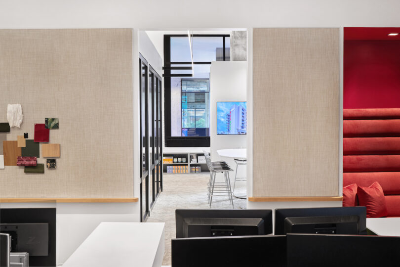 Modern office interior with open white desks, black monitors, and a red seating area in the background, visible through the doorway