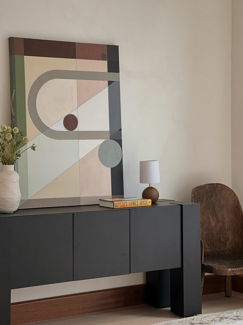 Modern living room with an abstract painting above a black sideboard that displays books and a lamp, next to a minimalist armchair.