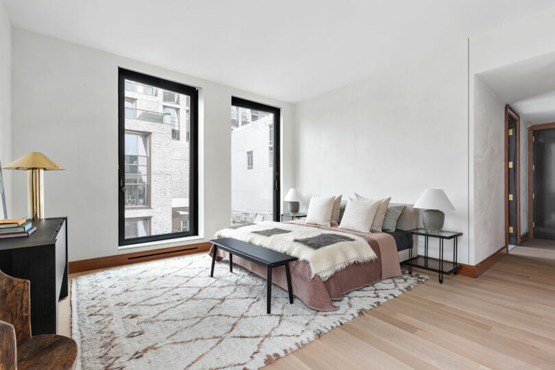 Modern bedroom with large windows, a plush bed with pillows, a bench at the foot, a patterned rug, and minimal décor.