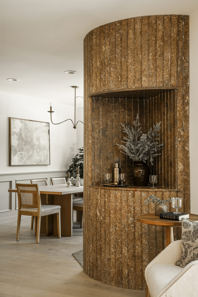 A rustic curved wooden wall featuring built-in shelving decorated with vases and plants, adjacent to a dining table and chairs.