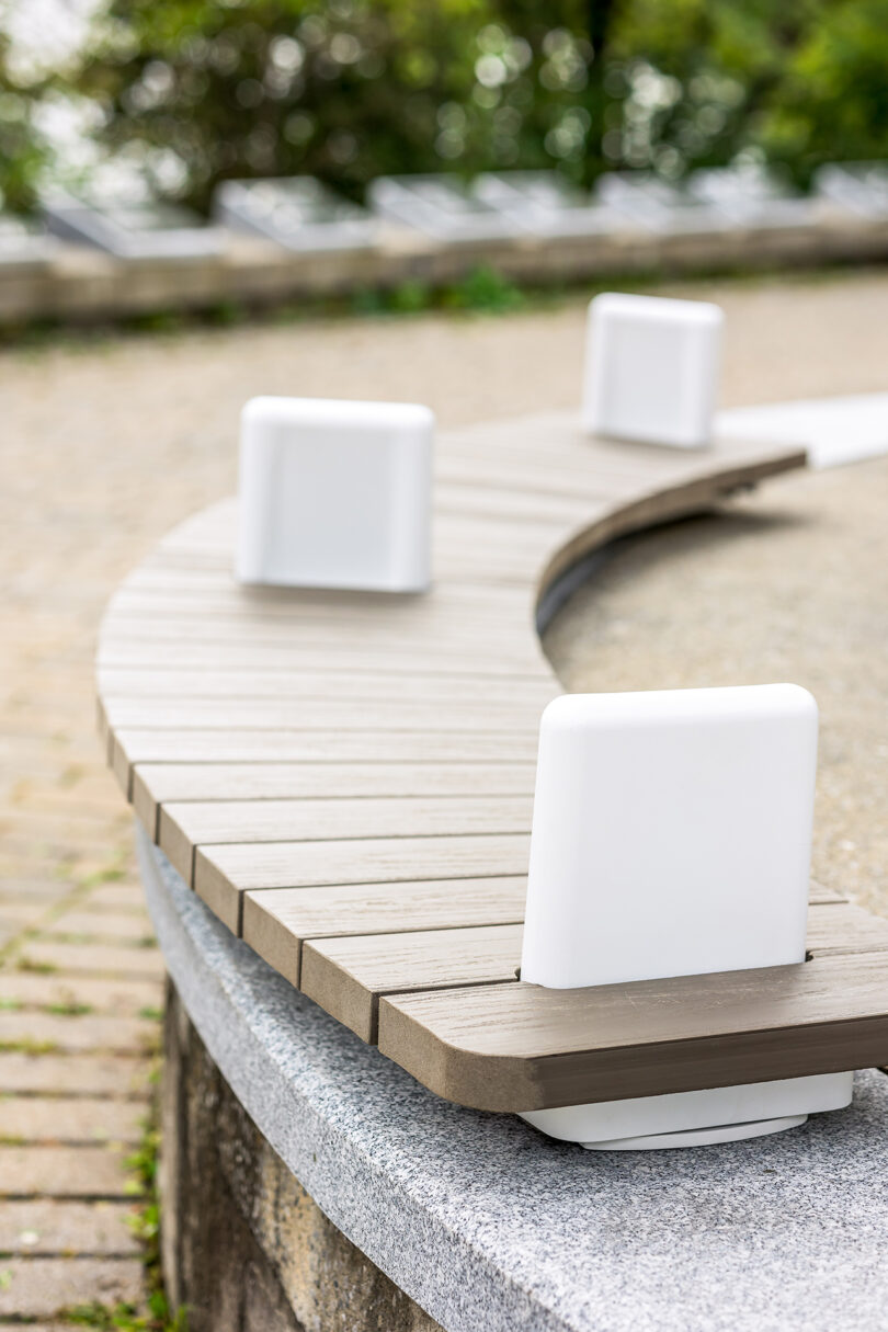 Detail of a curved wooden bench with white metal supports in a park with green grass and a path.