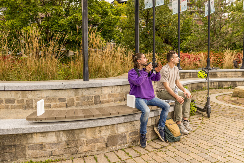 Two men sitting on a bench outdoors with backpacks beside them and greenery in the background.