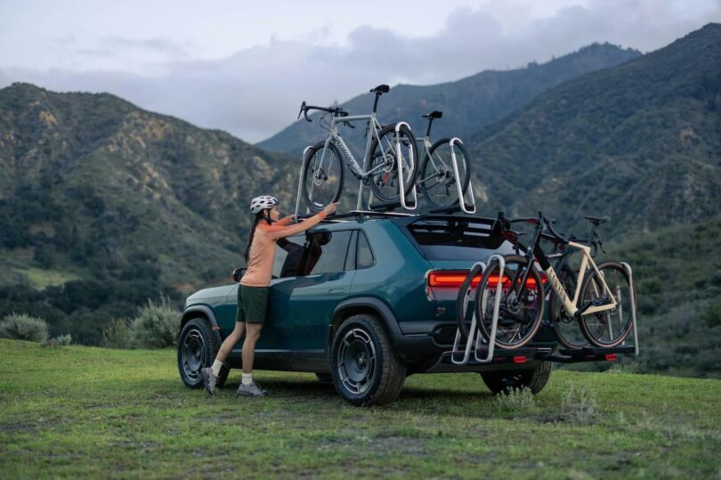 A person in a helmet loading bicycles onto a rack attached to the back of an suv in a mountainous area at dusk