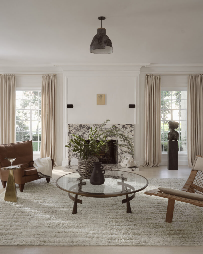 Living room with white walls, large windows, a marble fireplace, plush rug, and modern furniture, including a glass table and leather chairs.