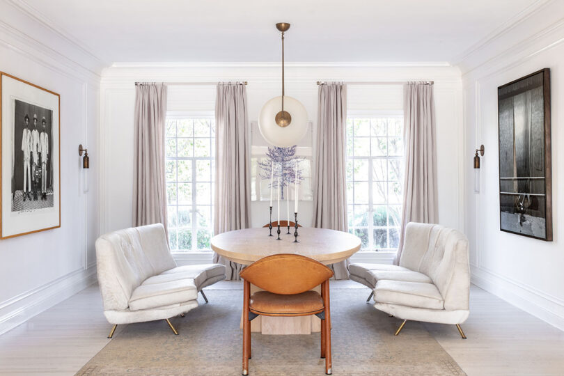 Dining room with a round wooden table, four chairs, large windows with curtains, and two large black-and-white photos on the walls.