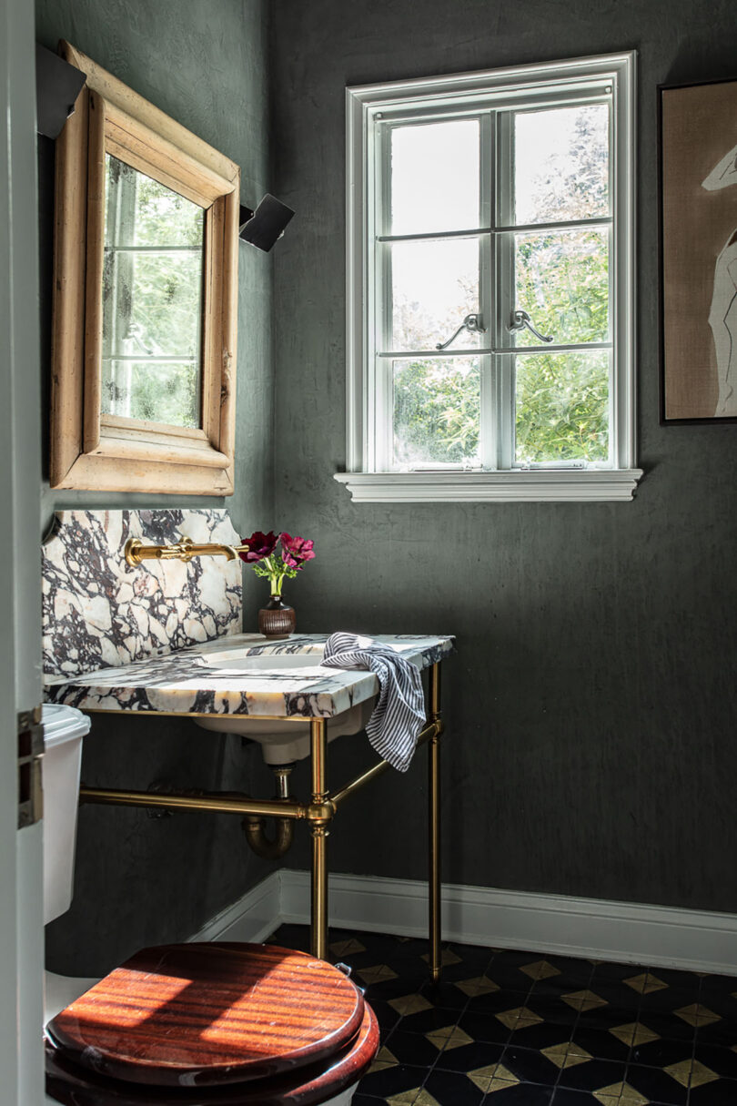 Bathroom with dark gray walls, a marble-top sink, gold fixtures and a rustic wooden framed mirror, and a small round table with a red stool near the window.