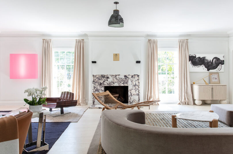 Living room featuring a marble fireplace, modern art on walls, curved sofa, wooden chairs, and large windows with beige drapes.