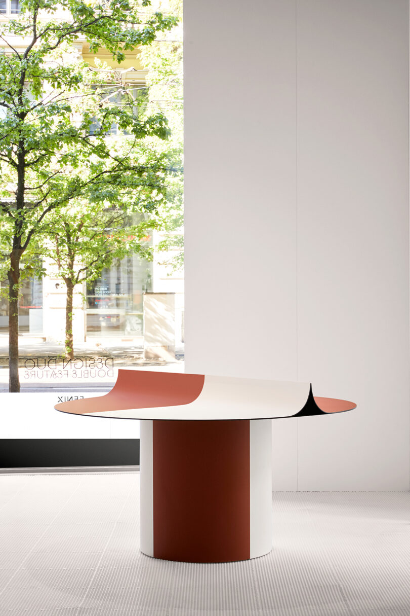 Curved table in shades of beige and terracotta with a central fold and a view of trees through large windows.
