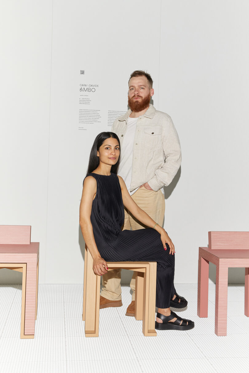 A man and a woman posing in an art gallery with minimalist furniture, the man standing and the woman sitting on a stool.
