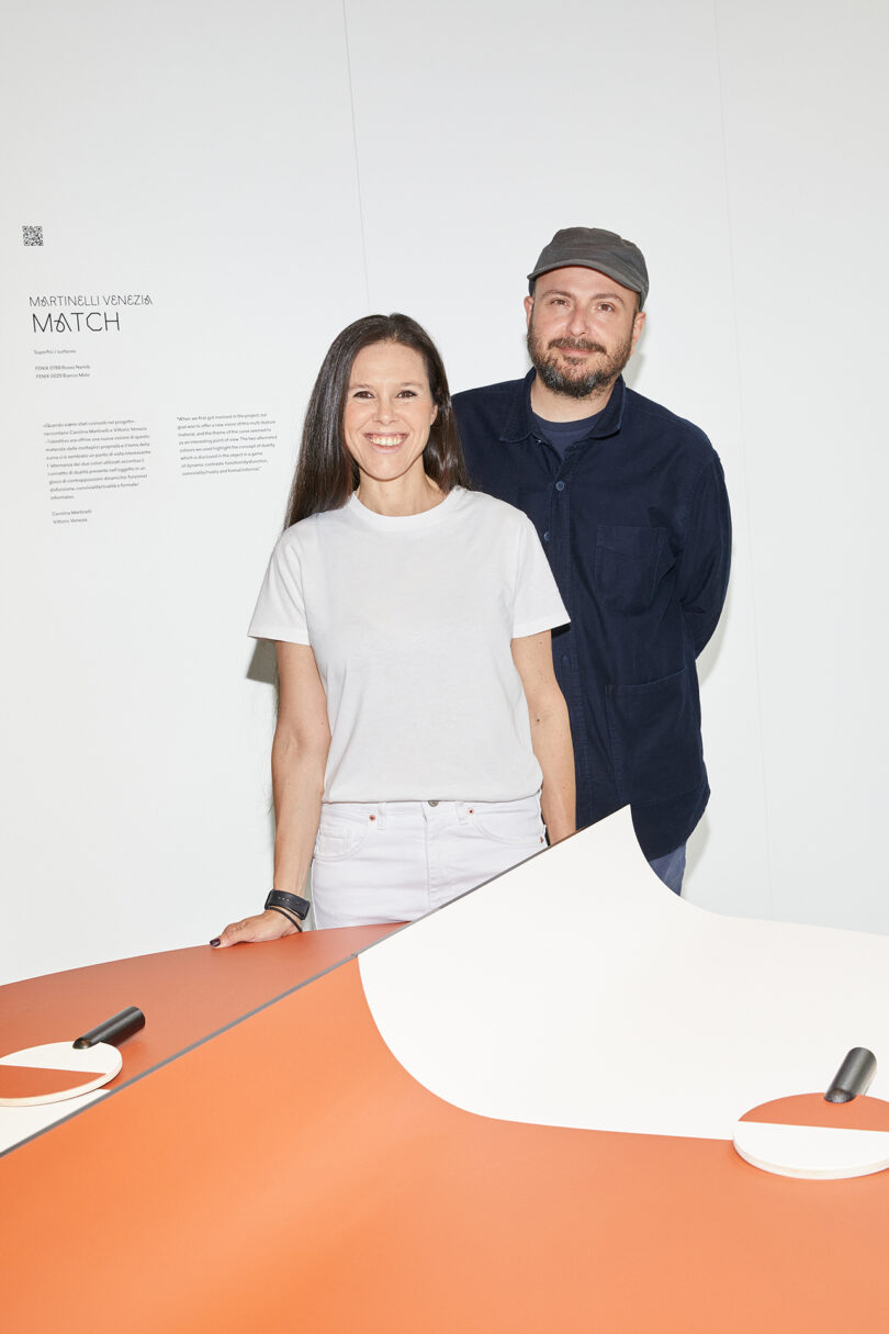 A man and a woman standing at a table in an art gallery, with informational text on the wall behind them.