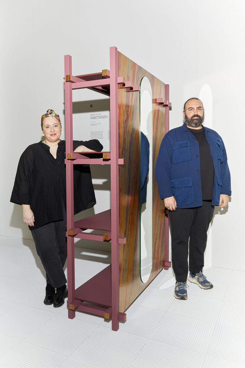 Two people standing beside a modern, multicolored shelving unit in a gallery setting. one wears a black outfit, the other in a blue jacket and jeans.