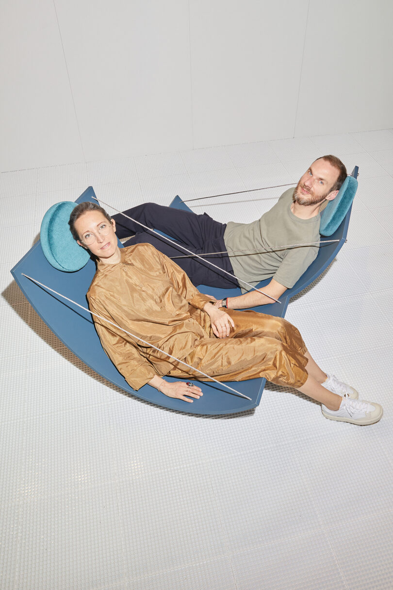 Two people lounging on a blue hammock in a gallery space, dressed in casual outfits, smiling at the camera.