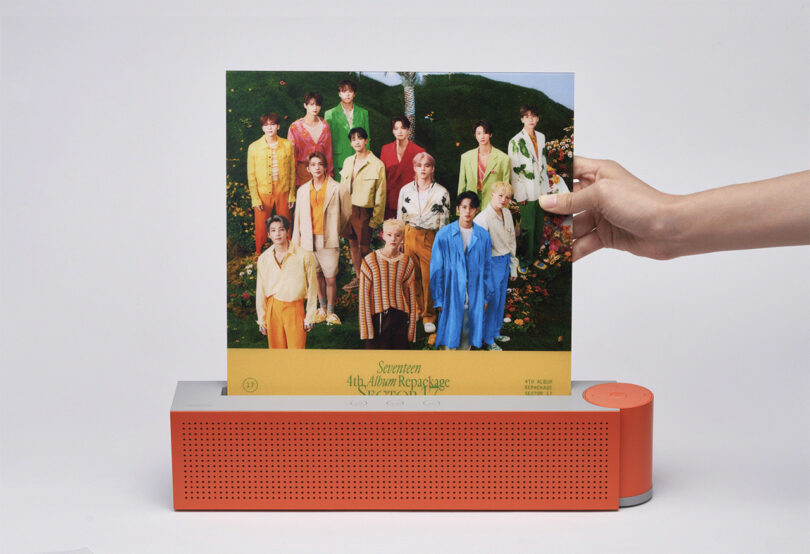 A hand holding up an album by a group of thirteen K-pop artists posing in colorful attire, displayed in a slot built into the orange-red Vibrary Digital LP Player.