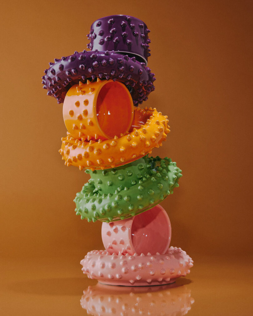 A stack of colorful textured mugs and saucers designed by Gustaf Westman, arranged vertically on a solid surface, against an orange background.