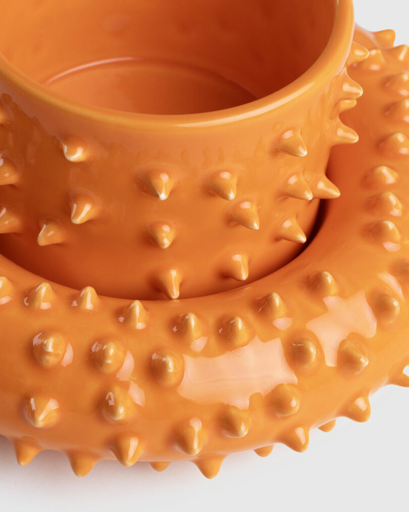 An orange ceramic Gustaf Westman mug and saucer with a textured, spiked surface stacked atop each other against a white background.