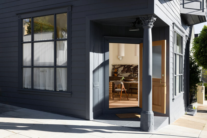 Exterior of a gray building with an open door revealing a cozy interior dining area, located on a sunny street corner.