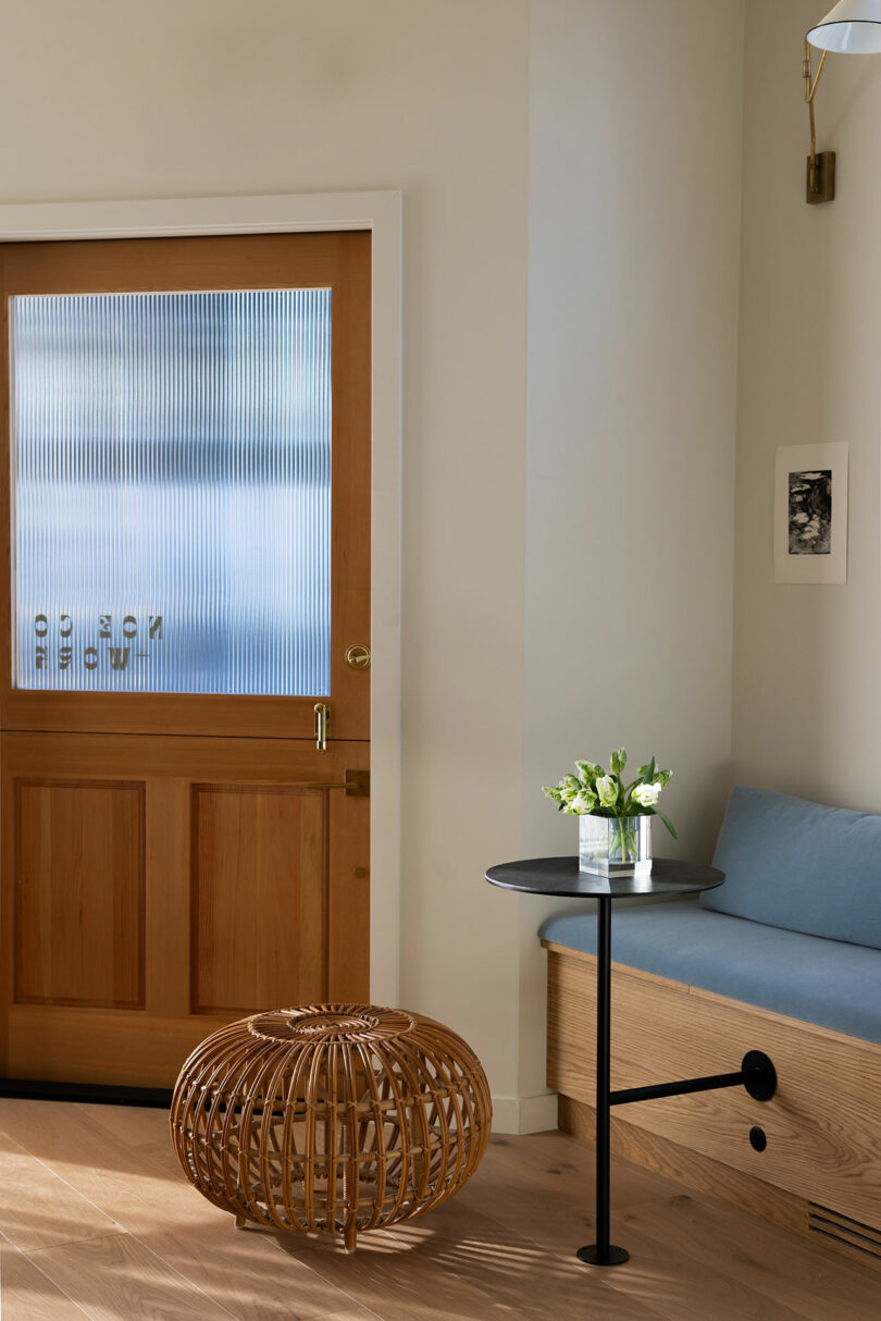 A modern entryway with a light blue bench, wooden door, black side table with a vase of flowers, and a woven ottoman.