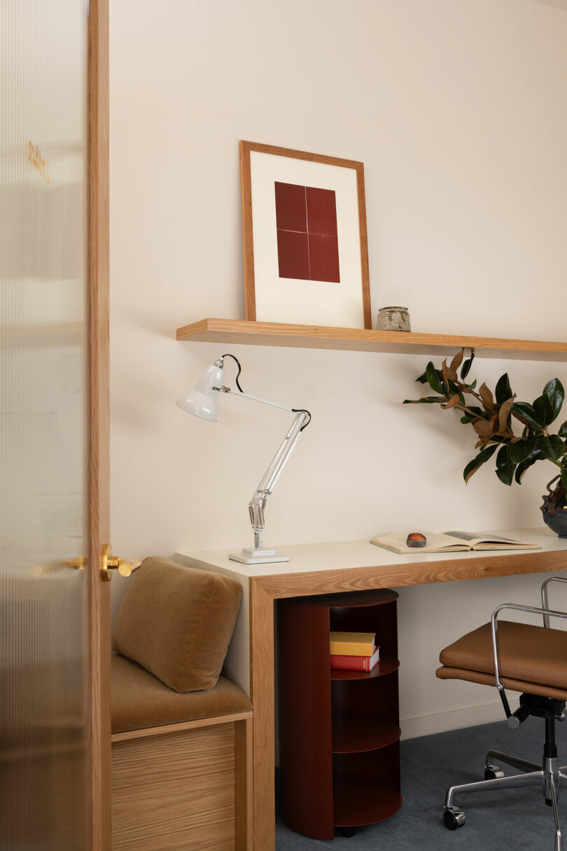 A cozy home office with a wooden desk, white adjustable lamp, framed artwork on the wall, and shelves with decorative items.