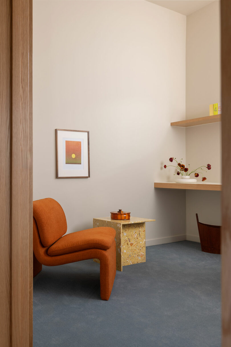 A minimalist room with a textured orange armchair, a golden decorative side table, artwork on the wall, and a shelf with vases.