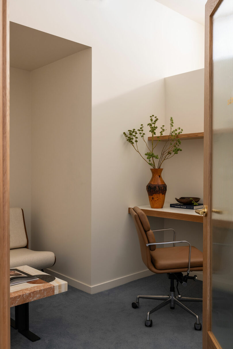 Small office space with a brown leather chair, wooden desk, beige carpet, and shelves with books and a vase with greenery.