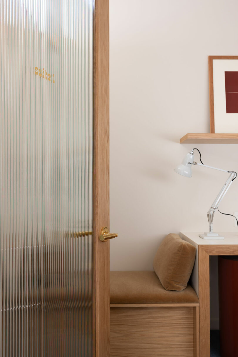 A small office space with a partially open wooden door showing frosted glass, next to a built-in desk and chair, with a white desk lamp turned on.