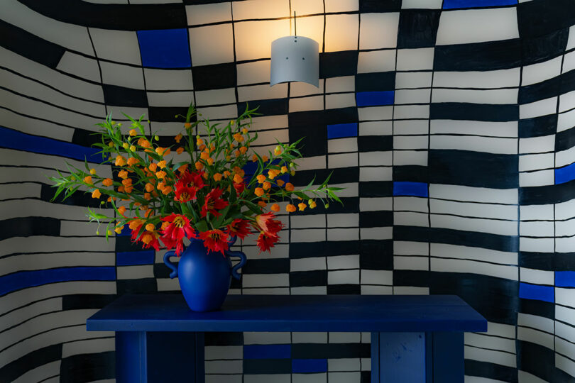 A bouquet of bright orange and red flowers in a blue vase on a blue table, set against a wavy black and white striped background.