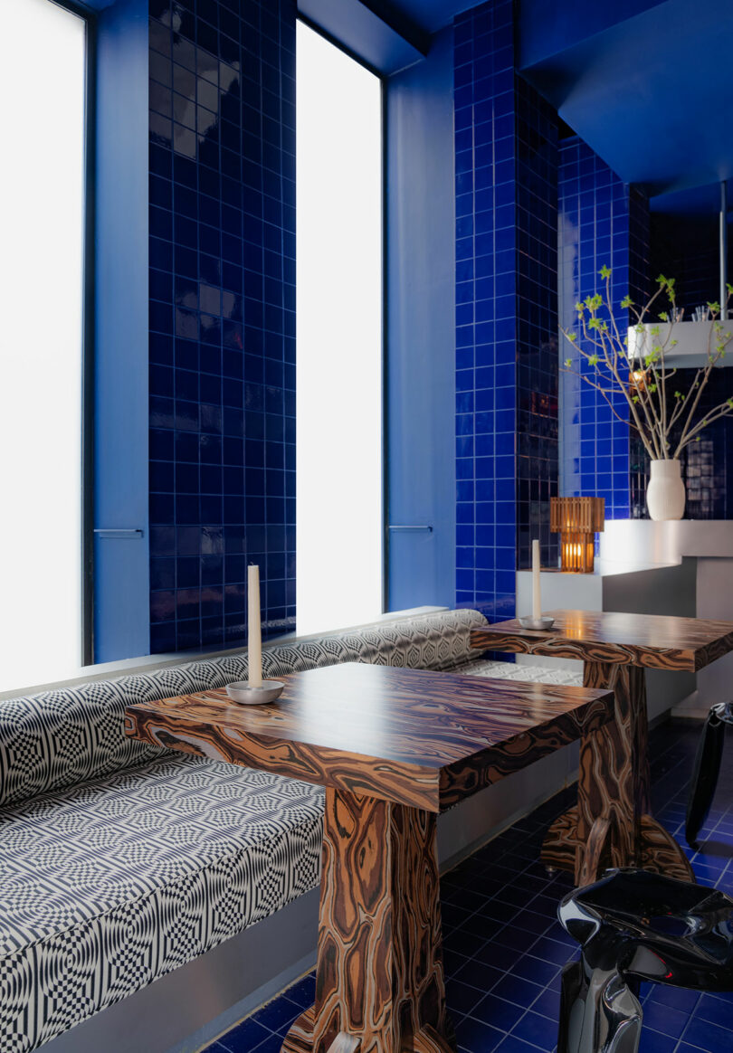 A modern dining area featuring blue tiled walls, a geometric bench cushion, wooden tables, and a white floor vase with yellow flowers next to large windows.