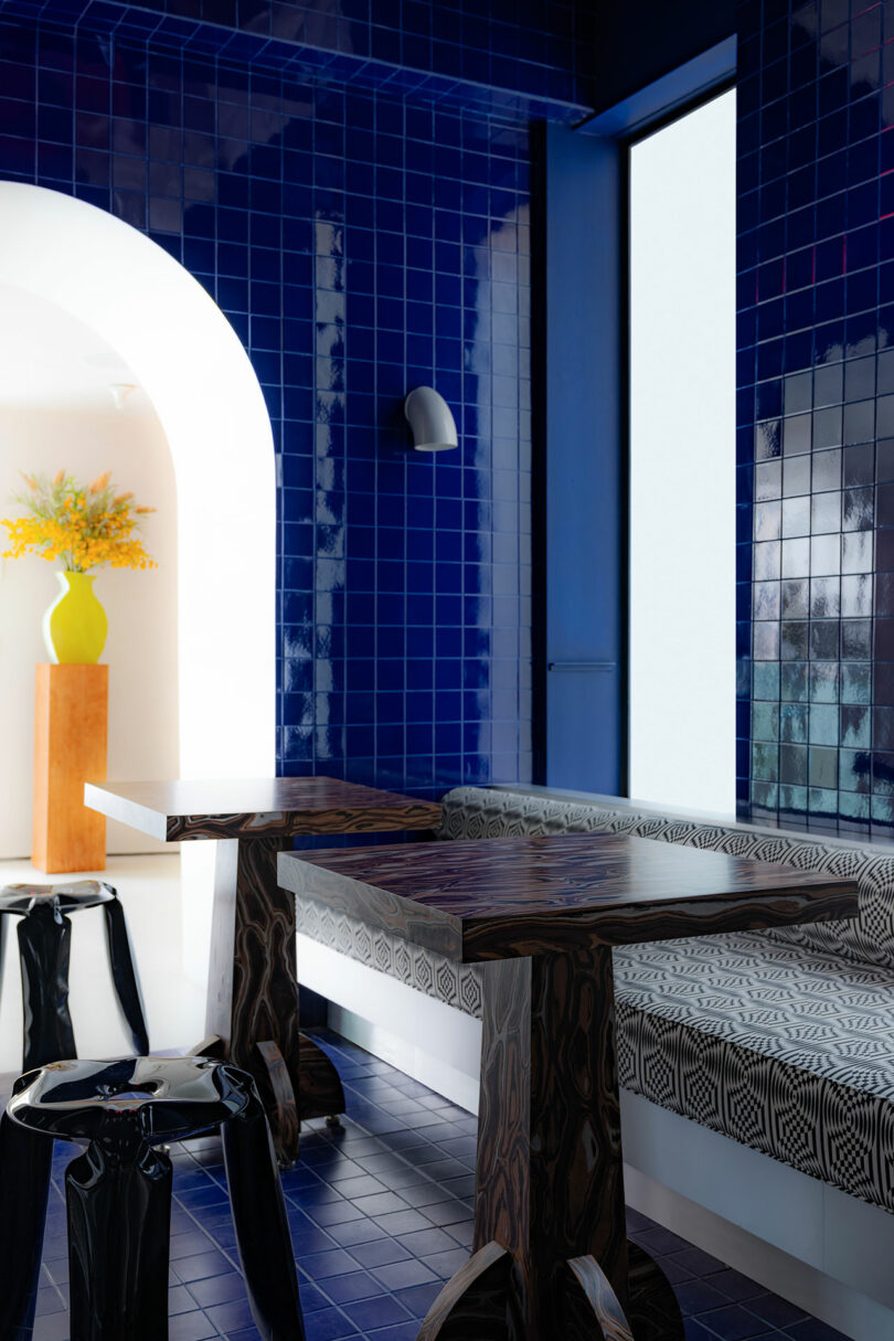 Interior view of a stylish lounge with a vibrant blue tiled wall, modern wooden tables, patterned benches, and a large arched doorway.