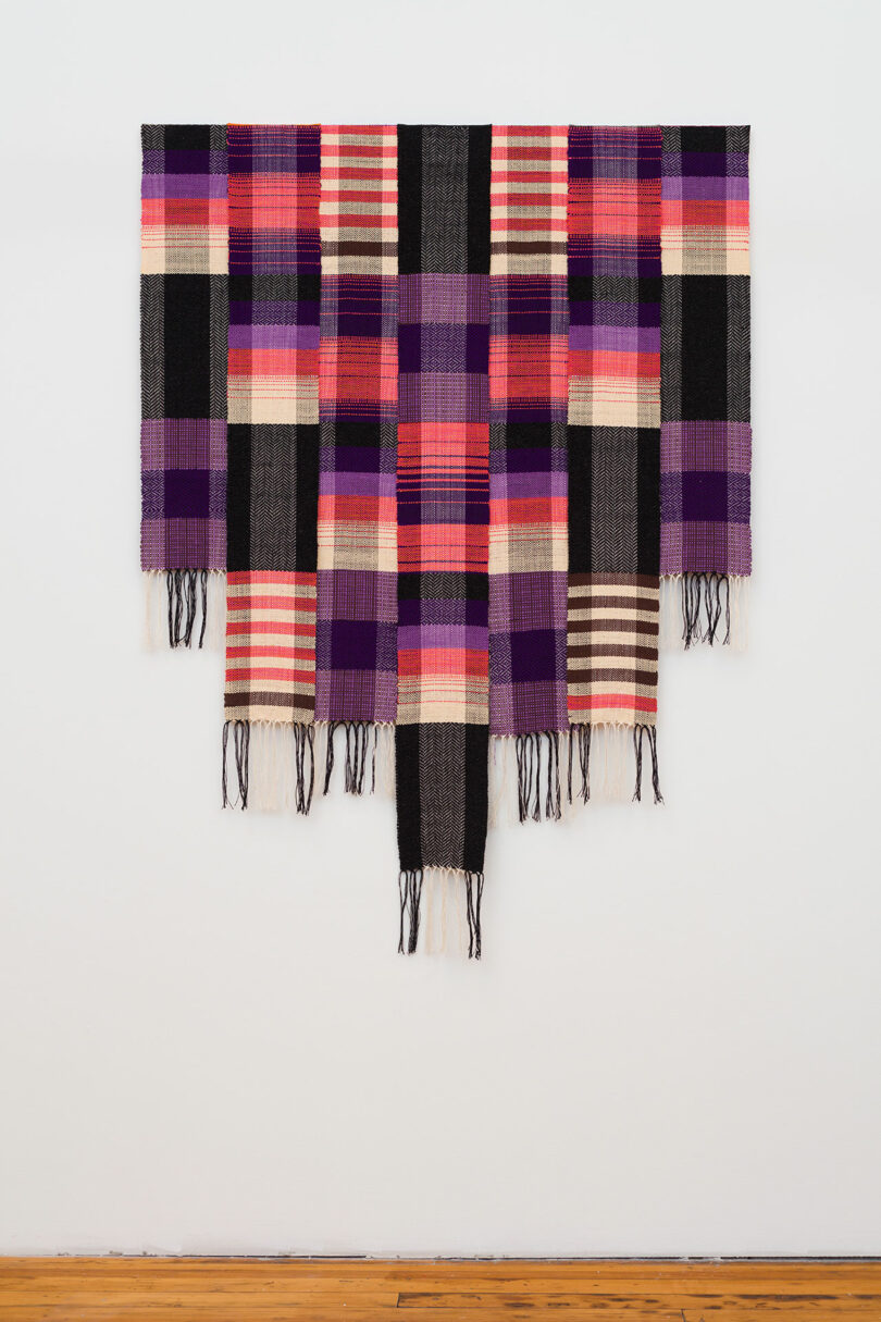 A large, multicolored woven tapestry with a plaid pattern hanging on a white wall, featuring primarily purple and red hues with fringed edges.