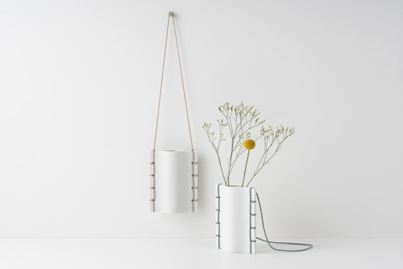 two white ceramic vases with simple foliage hanging on a white wall and on a white table