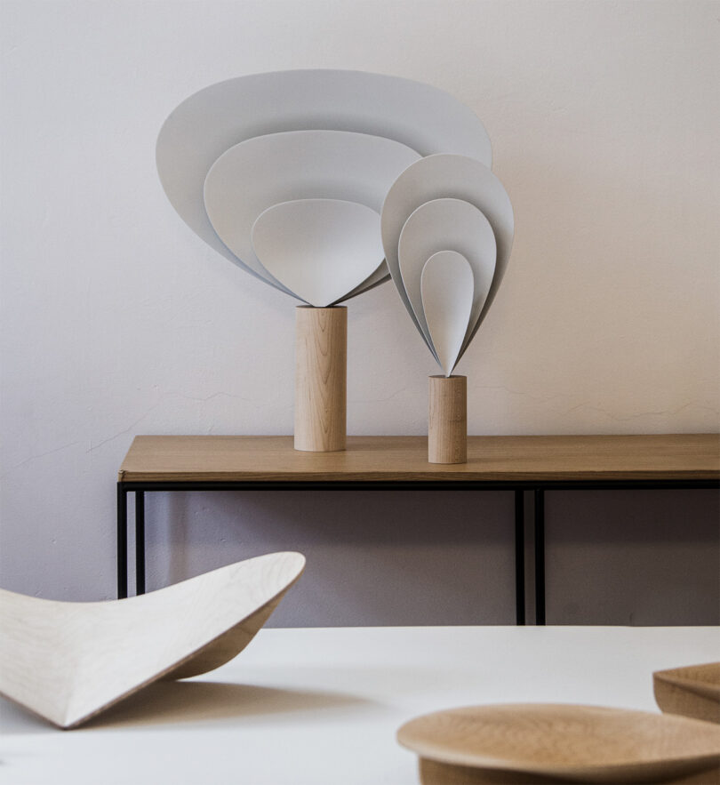 SWNA's Drop Light in small and large sizes set on shallow console table, each design comprised of three parabolic forms gathered into a fan-like shape nested into one another and held together by a cylindrical wood base. In foreground are a set of the Drop Trays celebrating Fritz Hansen's 150th anniversary.