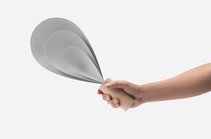 Person's hand holding a small Drop Light, a lamp design comprised of three parabolic forms gathered into a fan-like shape nested into one another and held together by a cylindrical wood base.