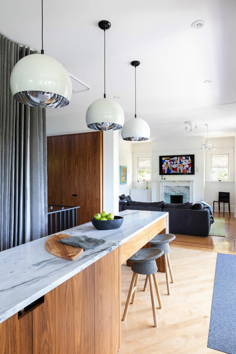 Modern kitchen interior featuring a marble bar counter, wooden cabinets, stylish pendant lights, and two bar stools near a window with sheer curtains.