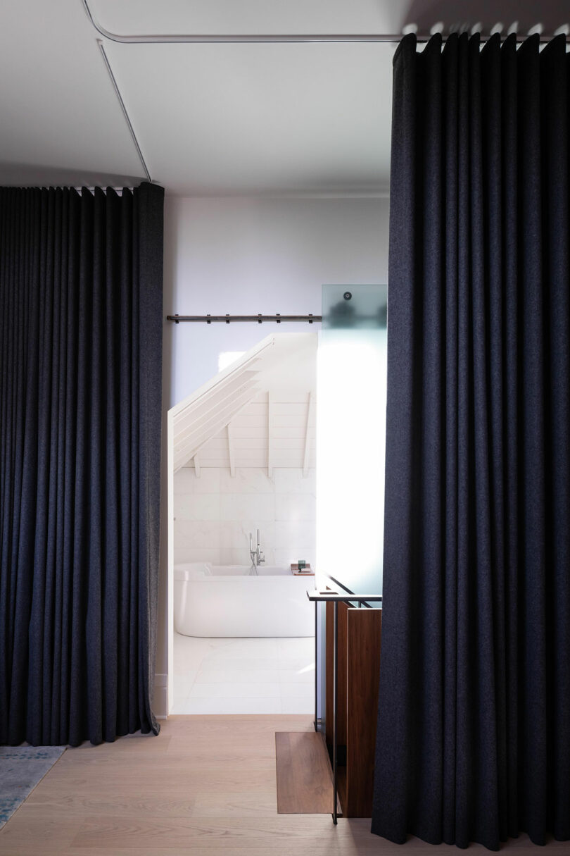 Modern bathroom viewed through dark curtains, featuring white marble walls, a visible bathtub, and wooden cabinet.