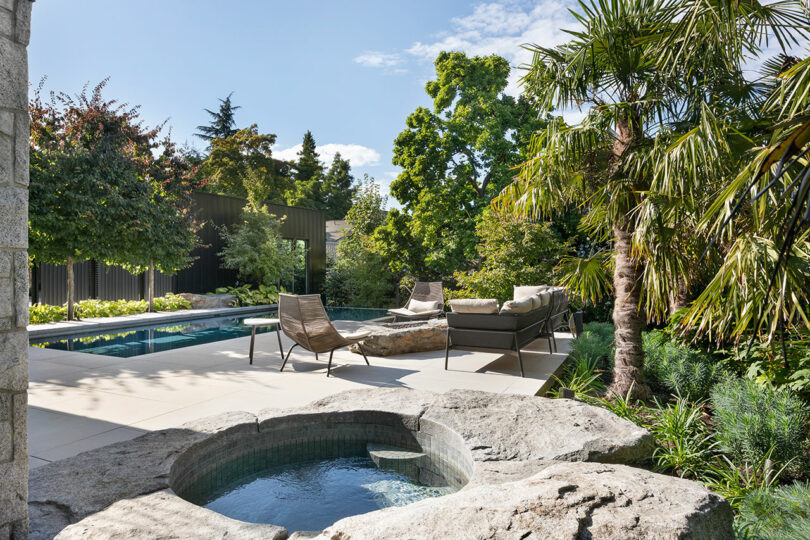 Lush garden with a small pool and jacuzzi, surrounded by tropical plants, stone features, and modern outdoor furniture near a larger swimming pool.