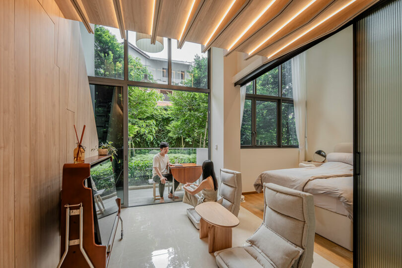 Modern studio apartment with wooden interiors, large windows overlooking greenery, and a young couple engaged in conversation at a small table.