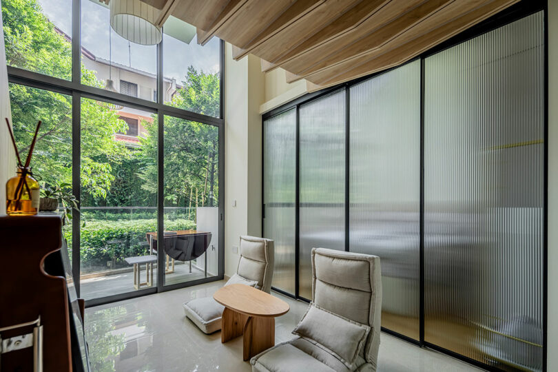 Modern living room featuring two cream chairs, a round wooden table, large glass windows with sheer curtains, and a view of a patio with outdoor furniture.