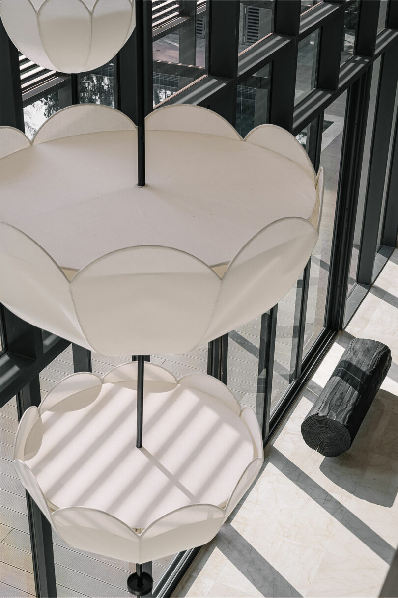 Modern interior with large, flower-shaped pendant lights and a wooden log bench by tall glass windows, casting soft shadows on a marble floor.