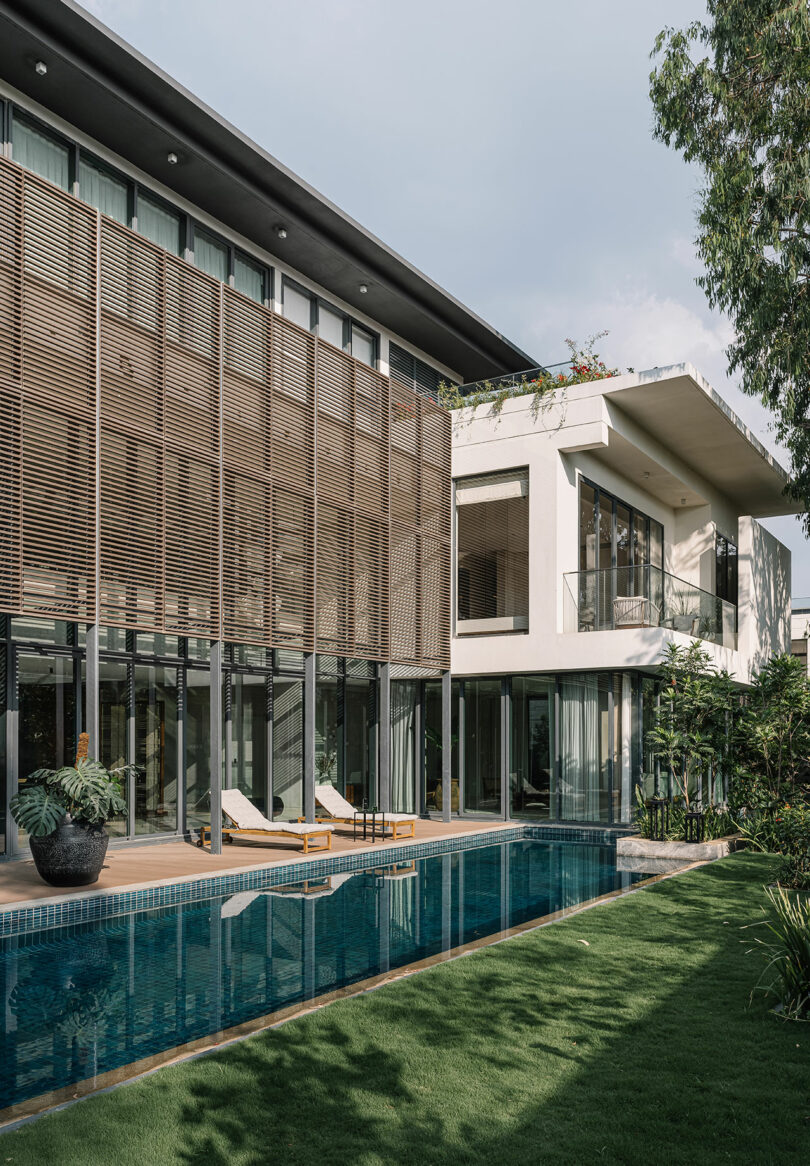 Modern two-story house with wooden slats, large windows, and a rectangular pool surrounded by a well-manicured lawn.