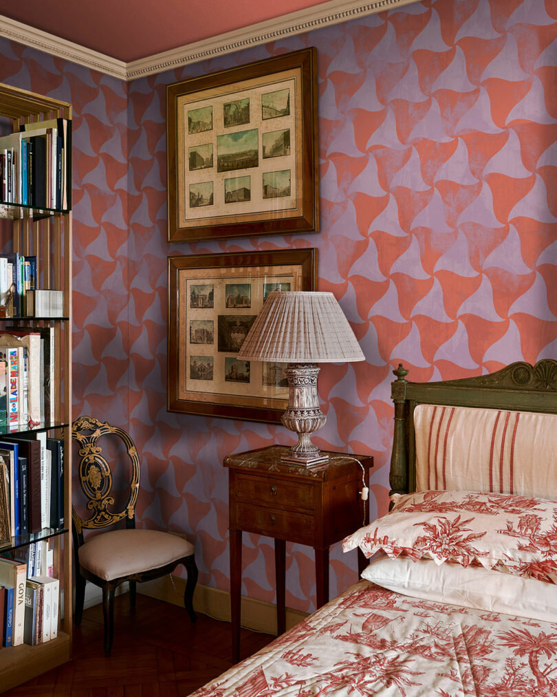 An eclectic bedroom with a patterned wallpaper, an antique wooden bed and side table, with framed artworks hanging on the wall.