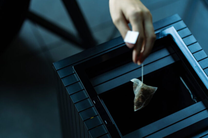 A person's hand placing a tea bag into a modern Zita-shaped black teapot on a dark surface.